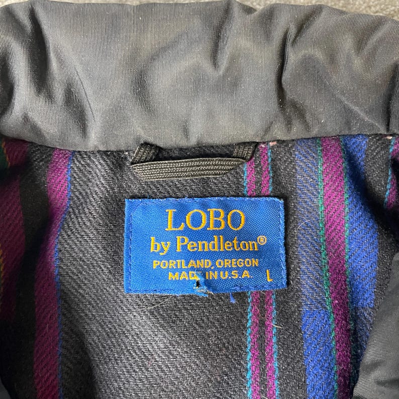 K&ouml;nnte beinhalten: Nahaufnahme eines Jackenkragens mit einem blauen Etikett mit der Aufschrift "LOBO by Pendleton" und "PORTLAND, OREGON MADE IN U.S.A.". Das Innenfutter hat ein farbenfrohes Streifenmuster. Die Jacke ist schwarz.