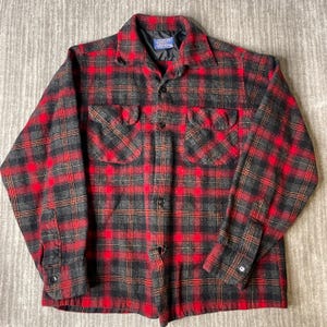 May include: A long-sleeve button-up shirt featuring a red and charcoal gray plaid pattern. The shirt includes a pointed collar, two chest pockets with buttoned flaps, and buttoned cuffs. A Pendleton brand label is visible at the inner neckline.