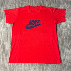 May include: A red t-shirt with the Nike logo in dark blue. The logo features the word "NIKE" above a curved checkmark. The shirt is made of a soft material and has short sleeves and a crew neck.