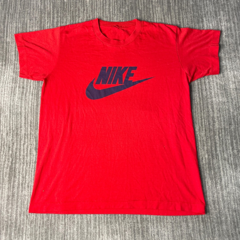May include: A red t-shirt with the Nike logo in dark blue. The logo features the word "NIKE" above a curved checkmark. The shirt is made of a soft material and has short sleeves and a crew neck.