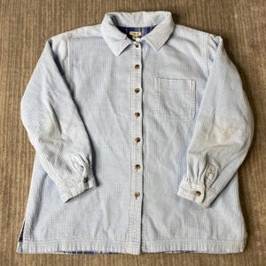 May include: A light blue corduroy button-down shirt featuring a classic collar, a single chest pocket, and long sleeves with buttoned cuffs. The shirt has a visible L.L.Bean label at the inner collar and a blue plaid lining visible at the hem.