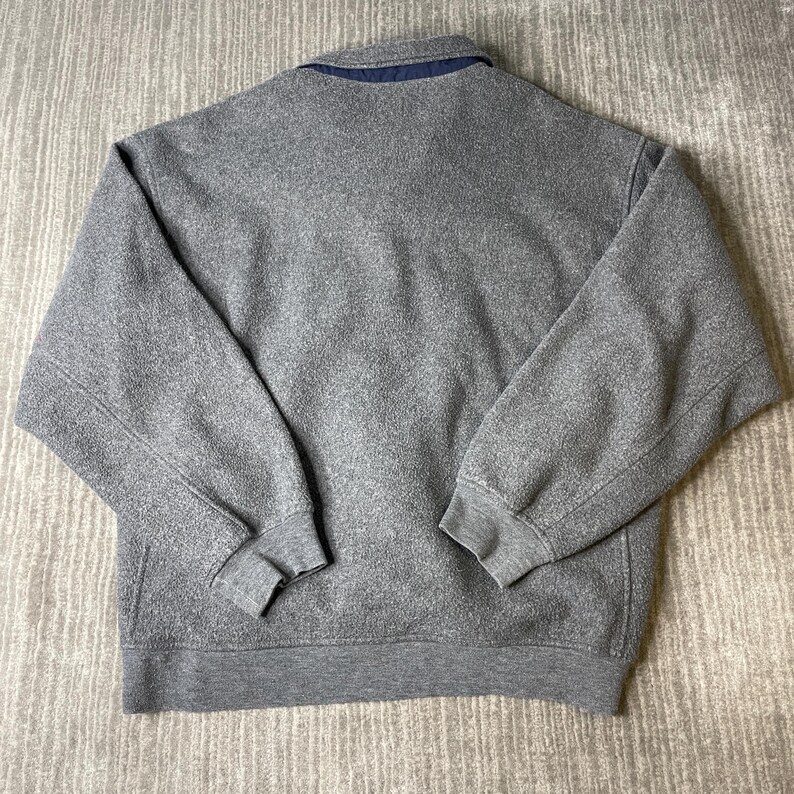 May include: A gray fleece sweatshirt with a collared neckline and a navy blue interior. The long-sleeved top has ribbed cuffs and a ribbed hem. The back of the sweatshirt is visible.
