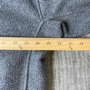 May include: Close-up of a gray fabric with a textured surface. A wooden ruler is placed across the fabric, showing measurements from 21 to 28 inches. The fabric appears to be part of a garment.