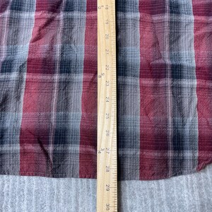 Op de afbeelding: Een close-up van een rood, zwart en grijs geruite stof. Een houten liniaal met zwarte markeringen en cijfers van 45,7 cm tot 76,2 cm is verticaal aan de rechterkant van de stof geplaatst. De stof heeft een getextureerde uitstraling.