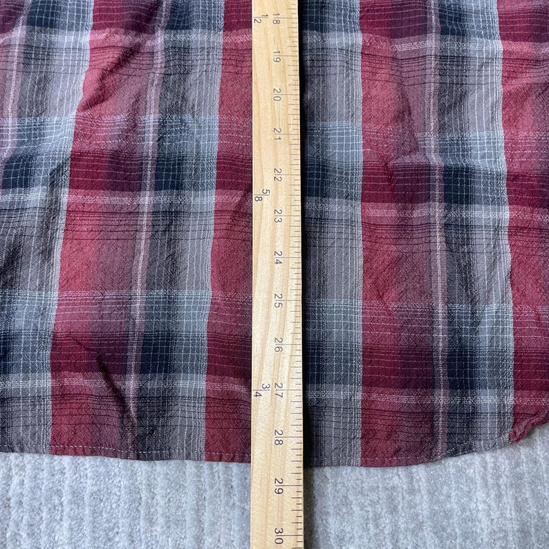 Op de afbeelding: Een close-up van een rood, zwart en grijs geruite stof. Een houten liniaal met zwarte markeringen en cijfers van 45,7 cm tot 76,2 cm is verticaal aan de rechterkant van de stof geplaatst. De stof heeft een getextureerde uitstraling.