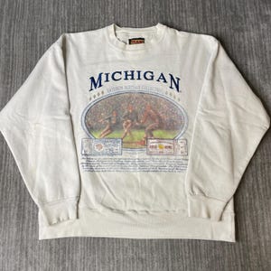 Könnte beinhalten: Ein cremefarbenes Langarm-Sweatshirt mit einem Grafikdruck und dem Text MICHIGAN GRIDIRON HERITAGE COLLECTION. Das Design enthält eine ovale Illustration von Footballspielern, zwei Vintage-Eintrittskarten und beschreibenden Text.