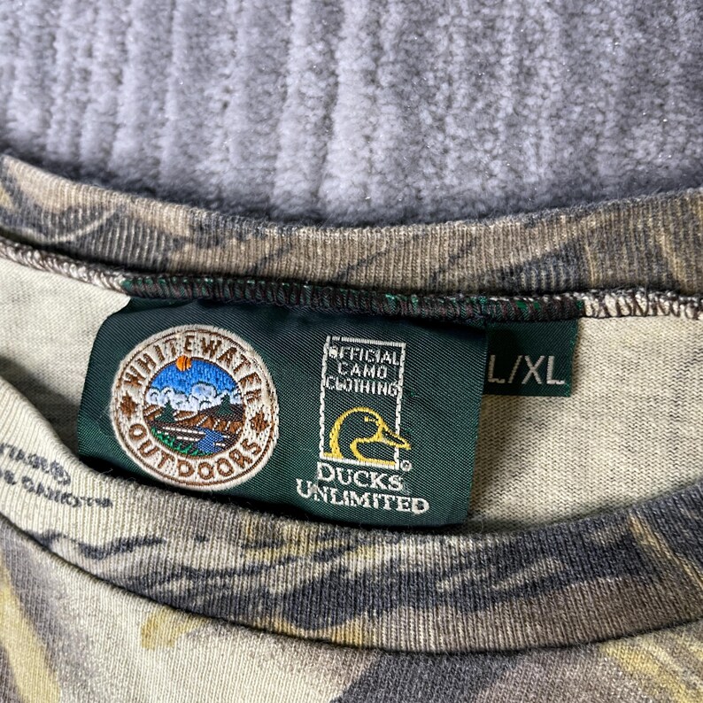 K&ouml;nnte beinhalten: Ein Hemd mit Tarnmuster und einem dunkelgr&uuml;nen Innenetikett am Kragen. Das Etikett zeigt das White Water Outdoors Logo, das offizielle Ducks Unlimited Camo Clothing Emblem mit einer gelben Entensilhouette und die Gr&ouml;&szlig;enangabe L/XL in Wei&szlig;.