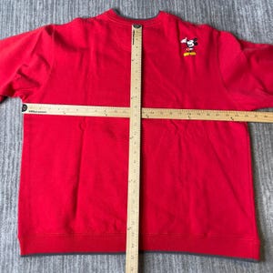 Puede incluir: Una sudadera roja de manga larga con una peque&ntilde;a figura bordada de Mickey Mouse en el pecho derecho. Dos reglas de madera est&aacute;n colocadas sobre la prenda, mostrando un ancho de 63,5 cm y una longitud vertical de 66 cm.
