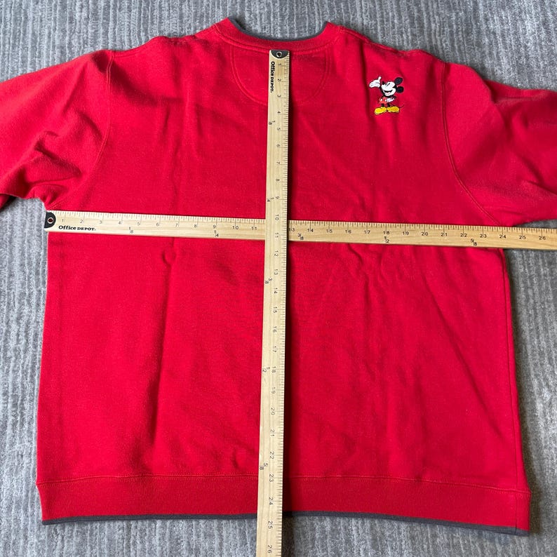 Puede incluir: Una sudadera roja de manga larga con una peque&ntilde;a figura bordada de Mickey Mouse en el pecho derecho. Dos reglas de madera est&aacute;n colocadas sobre la prenda, mostrando un ancho de 63,5 cm y una longitud vertical de 66 cm.