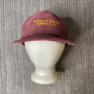 May include: A maroon corduroy baseball cap featuring the text Arizona State University embroidered in yellow thread on the front panel. The cap includes a matching maroon braided cord trim across the base of the crown above the brim.