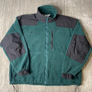 May include: A forest green fleece jacket featuring a high collar, a full-length front zipper, and black nylon overlays on the shoulders and elbows. The garment includes a vertical zippered chest pocket, elastic cuffs, and a drawstring hem.