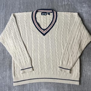 May include: A cream-colored cable knit sweater featuring a V-neckline with navy blue and red striped trim. The long-sleeved garment includes matching navy blue striped detailing on the cuffs and hem, with a label at the neck showing two sheep.