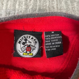Puede incluir: Una prenda roja con una etiqueta negra de Mickey & Co. que muestra un gr&aacute;fico circular de Mickey Mouse. Una etiqueta negra secundaria indica la talla M, 50% algod&oacute;n, 40% poli&eacute;ster, RN# 55219, Made in China y See Reverse for Care.