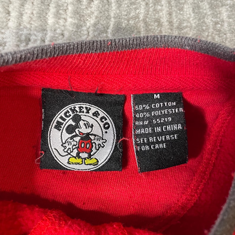 Puede incluir: Una prenda roja con una etiqueta negra de Mickey & Co. que muestra un gr&aacute;fico circular de Mickey Mouse. Una etiqueta negra secundaria indica la talla M, 50% algod&oacute;n, 40% poli&eacute;ster, RN# 55219, Made in China y See Reverse for Care.