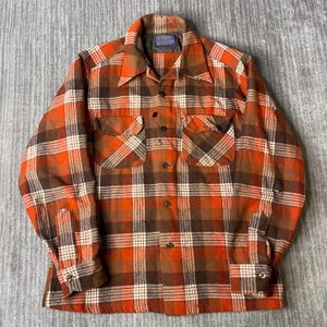 Peut inclure: Une chemise à manches longues boutonnée avec un motif à carreaux dans des tons orange, marron et blanc. La chemise présente un col pointu, deux poches de poitrine à rabat boutonné et des poignets boutonnés. Une étiquette Pendleton est visible.