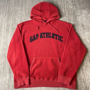 May include: A red fleece hooded sweatshirt featuring long sleeves, a front kangaroo pocket, and a drawstring hood. The text GAP ATHLETIC is embroidered in dark navy blue across the chest in a curved, bold, sans-serif font style.