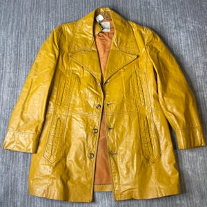 May include: A mustard yellow leather jacket featuring a wide notched collar, long sleeves, and a front button closure. The garment includes two vertical welt pockets on the chest and two lower pockets, all finished with visible stitching and a brown lining.