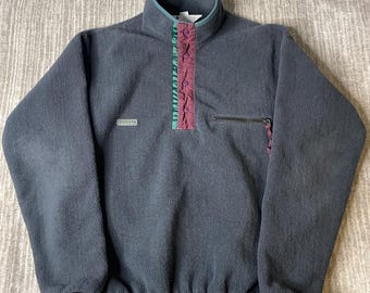 May include: A dark gray fleece pullover featuring a high collar with a teal and maroon snap-button placket. The garment includes a small chest pocket with a zipper closure and a rectangular brand logo patch on the right side of the chest.