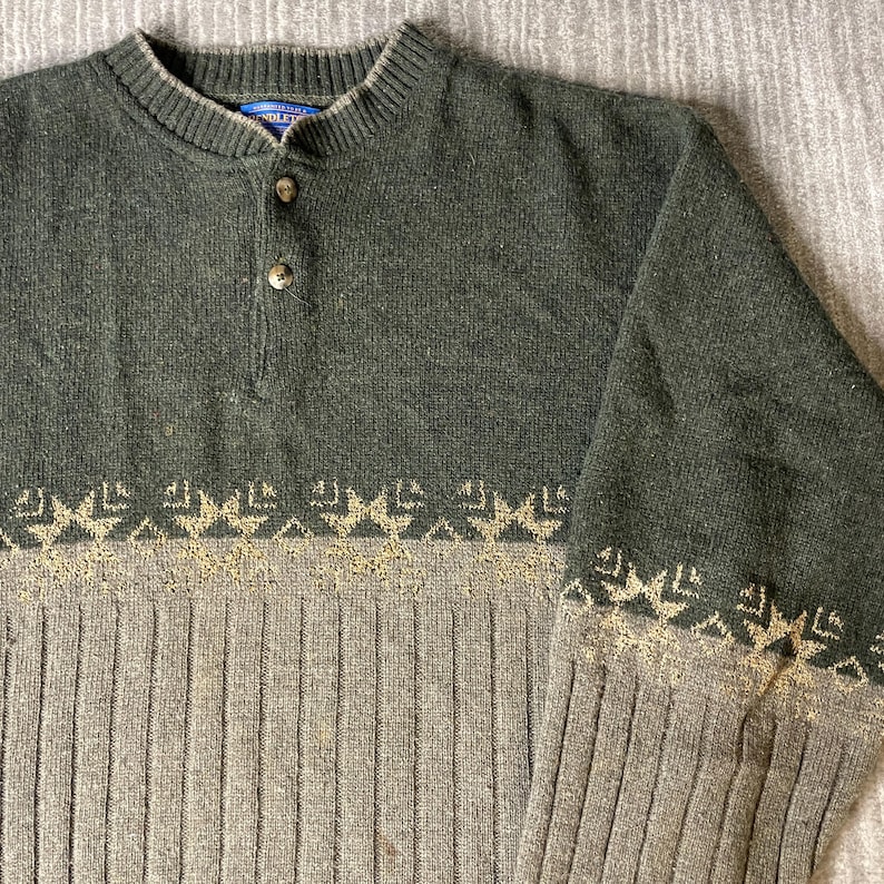 K&ouml;nnte beinhalten: Ein gr&uuml;ner und grauer Pullover mit Knopfkragen. Der obere Teil ist einfarbig gr&uuml;n, w&auml;hrend der untere Teil ein geripptes Design mit einem geometrischen Muster in einer helleren Farbe aufweist. Der Pullover hat einen klassischen, l&auml;ssigen Stil.