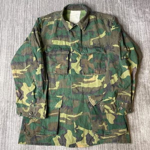 May include: A long-sleeve button-up shirt featuring a woodland camouflage pattern in shades of green, brown, and black. The shirt includes a pointed collar, two chest pockets with flaps, two lower front pockets with flaps, and buttoned cuffs.