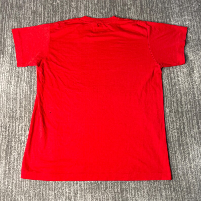 May include: A plain red t-shirt, seen from the back, lying flat on a grey surface. The short-sleeved shirt is made of a soft material and has a crew neck. The shirt is a vibrant, saturated red colour.