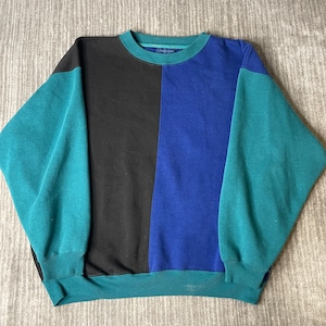 May include: A long-sleeve crewneck sweatshirt featuring a color-blocked design. The garment is divided into vertical panels of black and royal blue, with teal sleeves, a teal ribbed collar, and a teal ribbed hemline at the bottom of the torso.