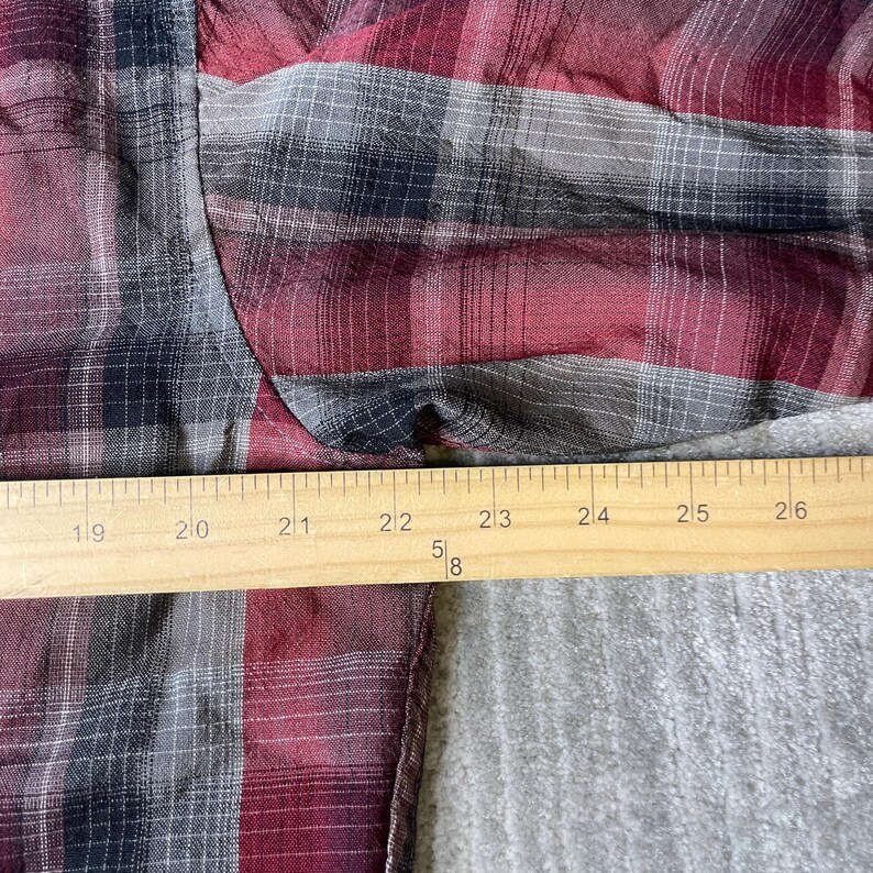 Op de afbeelding: Een close-up van een rood, zwart en grijs geruit overhemd. Een houten liniaal is over het overhemd geplaatst, metend van 48 tot 66 cm. De stof van het overhemd is gekreukeld en toont de textuur van het materiaal.