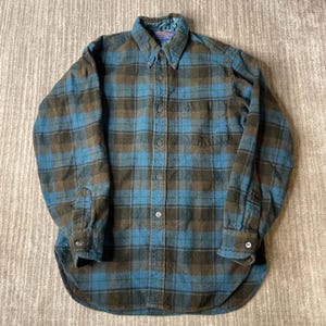 May include: A long-sleeve button-down shirt featuring a plaid pattern in shades of blue, brown, and dark green. The shirt has a pointed collar, a single chest pocket on the left side, buttoned cuffs, and a curved hemline with a visible brand label.