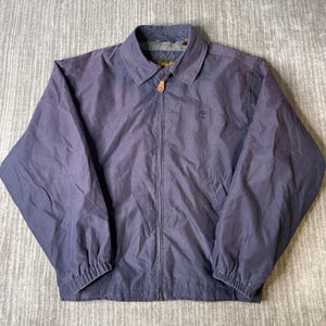 May include: A navy blue zip-up jacket featuring a classic collar, long sleeves with elastic cuffs, and a small embroidered Timberland tree logo on the left chest. The jacket includes a central zipper closure with a leather-like pull tab detail.