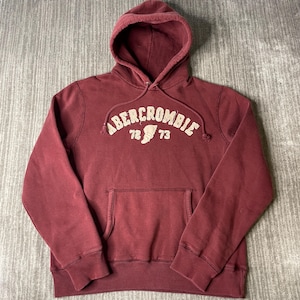 May include: A maroon long-sleeve hooded sweatshirt featuring a front kangaroo pocket and a drawstring hood. The chest displays the text ABERCROMBIE arched over the numbers 72 and 73 with a central graphic emblem in a light beige color.