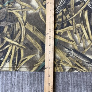 K&ouml;nnte beinhalten: Ein Stoff mit Camouflage-Muster in Braun-, Beige- und Dunkelgr&uuml;nt&ouml;nen, das trockenes Gras und Bl&auml;tter darstellt. Ein Holzlineal liegt vertikal auf dem Stoff und zeigt Messungen von 45 cm bis 74 cm entlang der Kante.