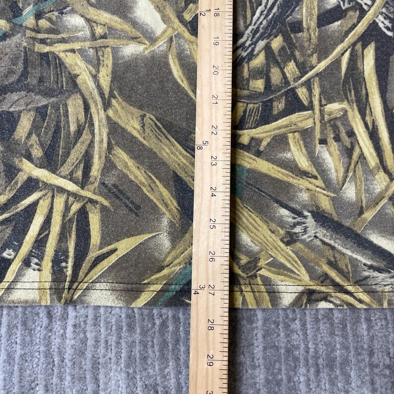 K&ouml;nnte beinhalten: Ein Stoff mit Camouflage-Muster in Braun-, Beige- und Dunkelgr&uuml;nt&ouml;nen, das trockenes Gras und Bl&auml;tter darstellt. Ein Holzlineal liegt vertikal auf dem Stoff und zeigt Messungen von 45 cm bis 74 cm entlang der Kante.