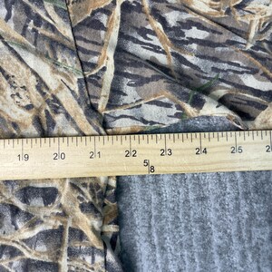 以下が含まれることがあります： 迷彩柄の生地と木製の定規のクローズアップ。生地は茶色、緑、ベージュの色合いで、自然なデザインを作り出しています。定規はセンチメートルでマークされています。