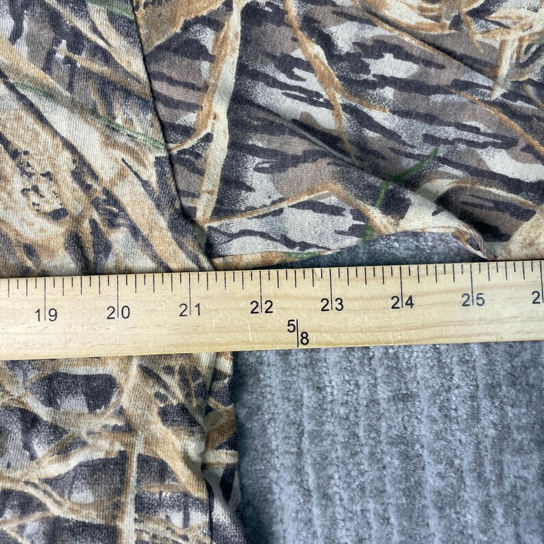 以下が含まれることがあります： 迷彩柄の生地と木製の定規のクローズアップ。生地は茶色、緑、ベージュの色合いで、自然なデザインを作り出しています。定規はセンチメートルでマークされています。