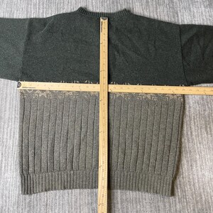 K&ouml;nnte beinhalten: Ein zweifarbiger Pullover mit Rippenmuster. Der obere Teil ist dunkelgr&uuml;n, w&auml;hrend der untere Teil hellgrau mit vertikalen Rippen ist. Ein Holzlineal liegt &uuml;ber dem Pullover und misst seine Abmessungen.