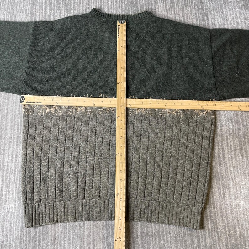 K&ouml;nnte beinhalten: Ein zweifarbiger Pullover mit Rippenmuster. Der obere Teil ist dunkelgr&uuml;n, w&auml;hrend der untere Teil hellgrau mit vertikalen Rippen ist. Ein Holzlineal liegt &uuml;ber dem Pullover und misst seine Abmessungen.