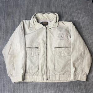 May include: A light beige jacket with a zip-up front, two front pockets, and a collared neckline. The jacket has a logo on the upper left chest and a small tag near the collar. The jacket is made of a lightweight material.
