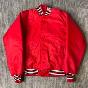May include: A bright red varsity jacket featuring a snap-button front closure. The collar, cuffs, and hem are finished with ribbed knit trim in alternating stripes of black, white, and red. The jacket has long sleeves and a smooth, satin-like finish.