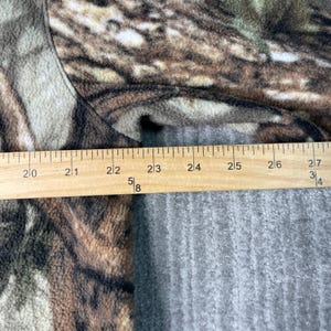 以下が含まれることがあります： 迷彩柄の生地のクローズアップ。定規が上に置かれています。定規は50.8 cmから68.6 cmを測定します。生地は茶色、緑、グレーの色合いです。