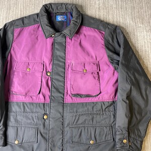 K&ouml;nnte beinhalten: Eine Vintage-Jacke mit Colorblock-Design. Die obere Brust und die Taschen sind in leuchtendem Lila gehalten, kontrastiert durch einen schwarzen Kragen und &Auml;rmel. Der untere Teil der Jacke ist dunkelgrau, mit vier Taschen und goldfarbenen Kn&ouml;pfen.