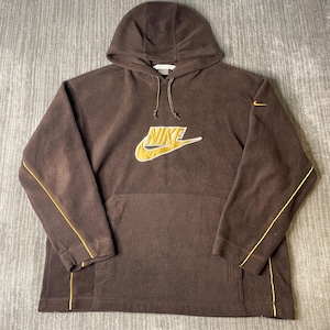 May include: Brown Nike hoodie with a gold and white logo on the front. The hoodie has a drawstring and gold trim along the sleeves. The Nike logo is embroidered with a gold outline and white fill.