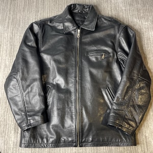 Könnte beinhalten: Eine schwarze Lederjacke mit durchgehendem Frontreißverschluss, klassischem Kragen, langen Ärmeln und einer Reißverschlusstasche auf der linken Brustseite. Die Jacke verfügt über zwei untere Seitentaschen und eine glatte Oberfläche.