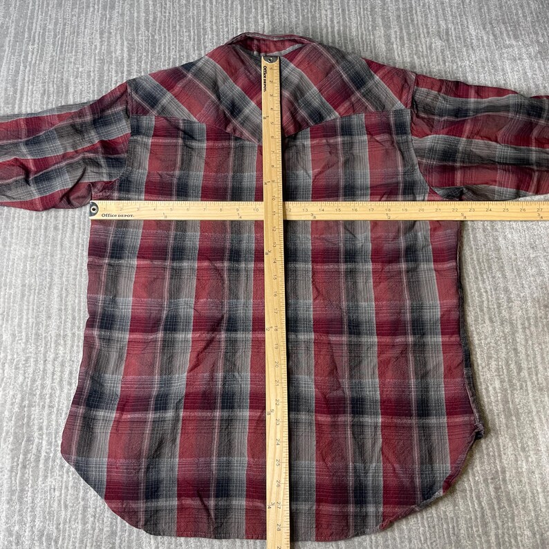 Op de afbeelding: Een rood, zwart en grijs geruit button-down shirt. Het shirt wordt gemeten met een houten liniaal. Het shirt heeft lange mouwen en een klassieke kraag. Het shirt is gemaakt van een zachte, geweven stof.