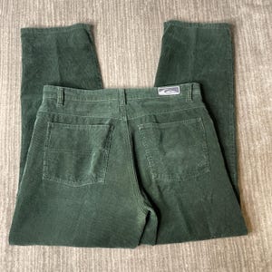 May include: Dark forest green corduroy pants featuring a straight-leg cut, a button and zipper fly closure, belt loops, and two back patch pockets. A rectangular grey and white brand label is stitched above the right back pocket.