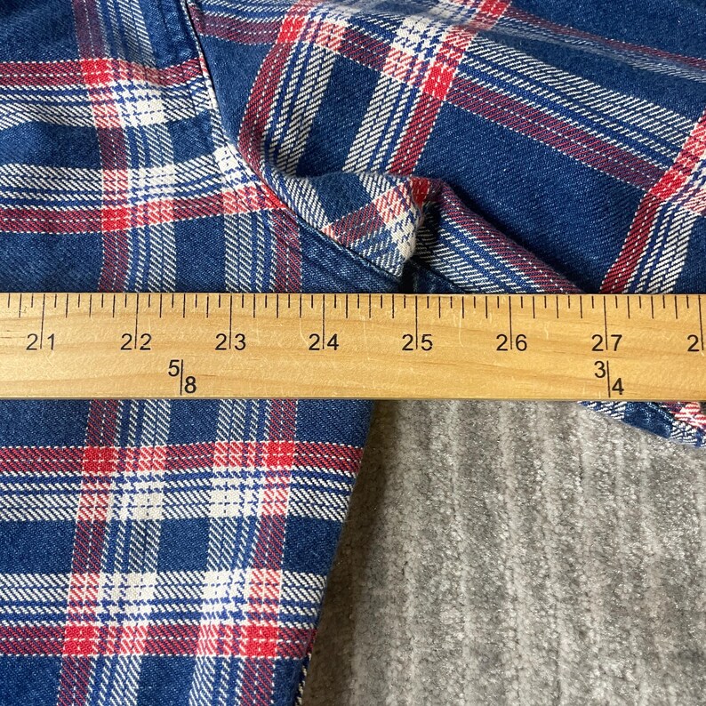 May include: Close-up of a blue, red, and white plaid shirt. A wooden ruler is placed across the fabric, measuring approximately 6 inches. The shirt's pattern is a classic design, suitable for casual wear.