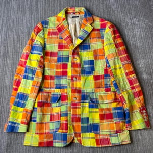 May include: A long-sleeve blazer featuring a vibrant patchwork design composed of various plaid fabric squares in bright shades of yellow, orange, red, blue, and green. The garment includes a notched lapel, three front buttons, and two pockets.