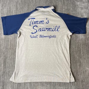 May include: A short-sleeve polo shirt with a light gray body and dark blue sleeves and collar. The back features blue embroidered text that reads Timm's Sawmill and West Bloomfield in a cursive script style across the upper back area.