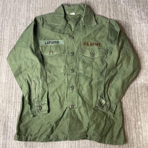 May include: An olive green long-sleeve button-up shirt featuring a pointed collar, two chest pockets with button flaps, and buttoned cuffs. The shirt displays a name patch reading LAPAKKO and a U.S. ARMY patch above the chest pockets.