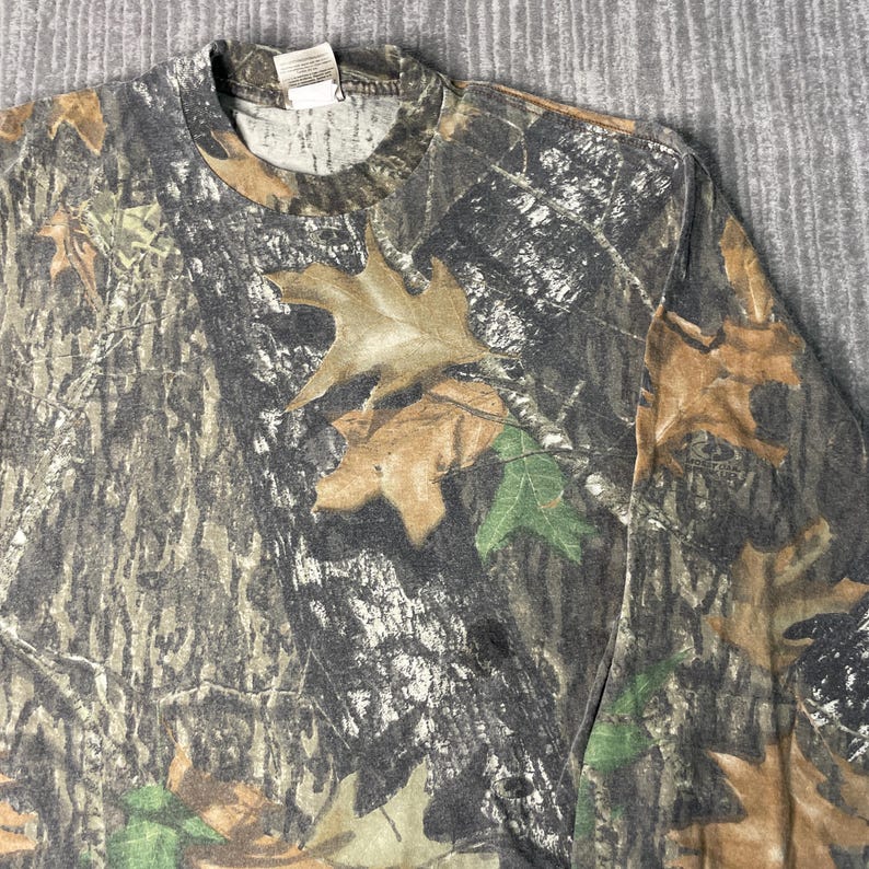 May include: Long-sleeved camouflage shirt featuring a pattern of brown, green, and gray leaves and branches. The shirt has a crew neck and a tag at the back of the neck. The fabric appears to be a blend of cotton and polyester.