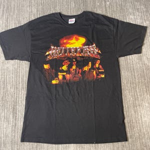 May include: A black short-sleeve t-shirt featuring a graphic print of five individuals sitting at a table with drinks. Above the group is a large orange flame and the text "HELLYEAH" in a metallic, jagged font. The shirt has a crew neckline.